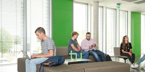 Sechs Studierende sitzen in einem Aufenthaltsbereich in einem Uni-Gebäude. Sie lesen Bücher, arbeiten am Laptop oder unterhalten sich.